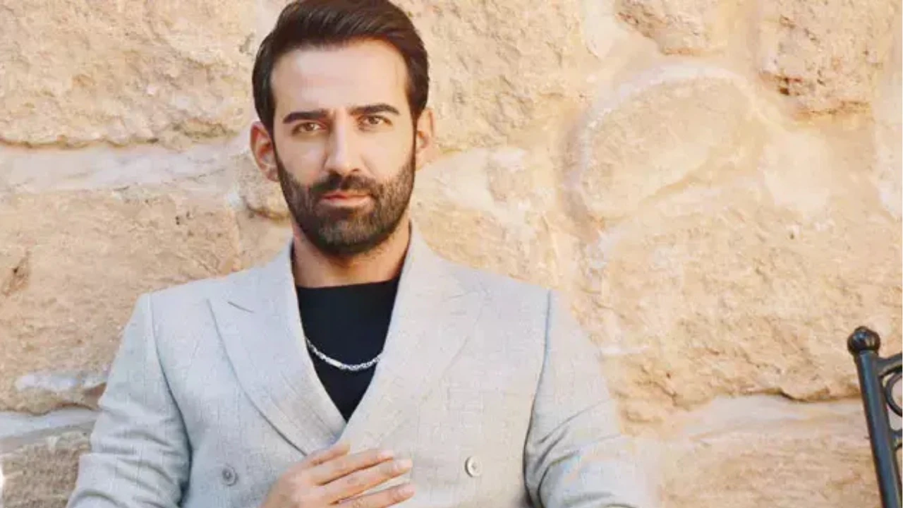 Ferit Kaya, açık gri takım ceket ve siyah tişört kombinini gümüş bir kolyeyle tamamlamış halde taş duvar önünde poz verirken. Bakımlı sakalı ve etkileyici bakışlarıyla profesyonel bir çekim için kameraya bakıyor. Ferit Kaya, açık gri takım ceket ve siyah tişört kombinini gümüş bir kolyeyle tamamlamış halde taş duvar önünde poz verirken. Bakımlı sakalı ve etkileyici bakışlarıyla profesyonel bir çekim için kameraya bakıyor.